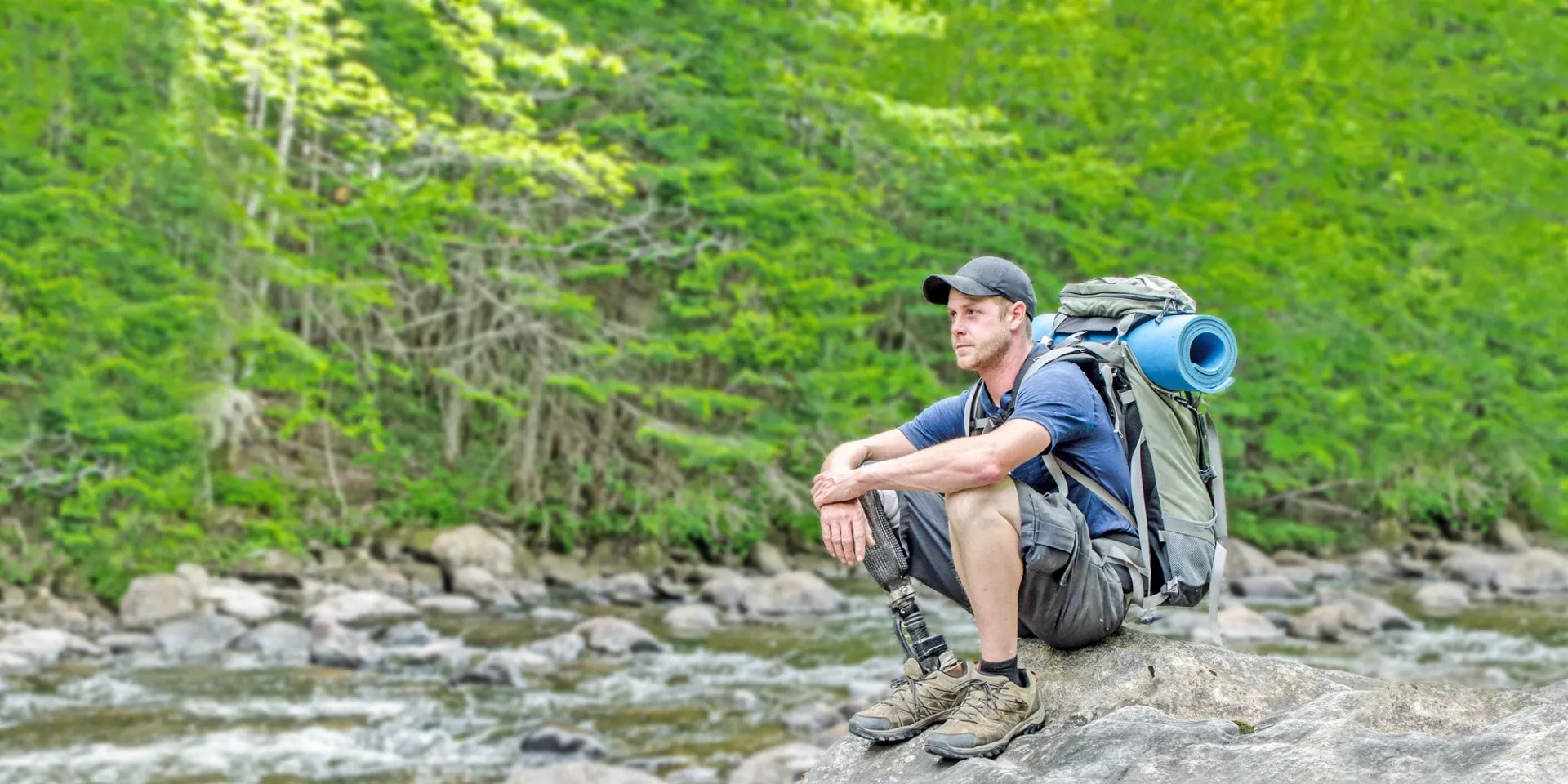 Man with prosthetic leg and hiking gear sitting on rock by river in forest setting.