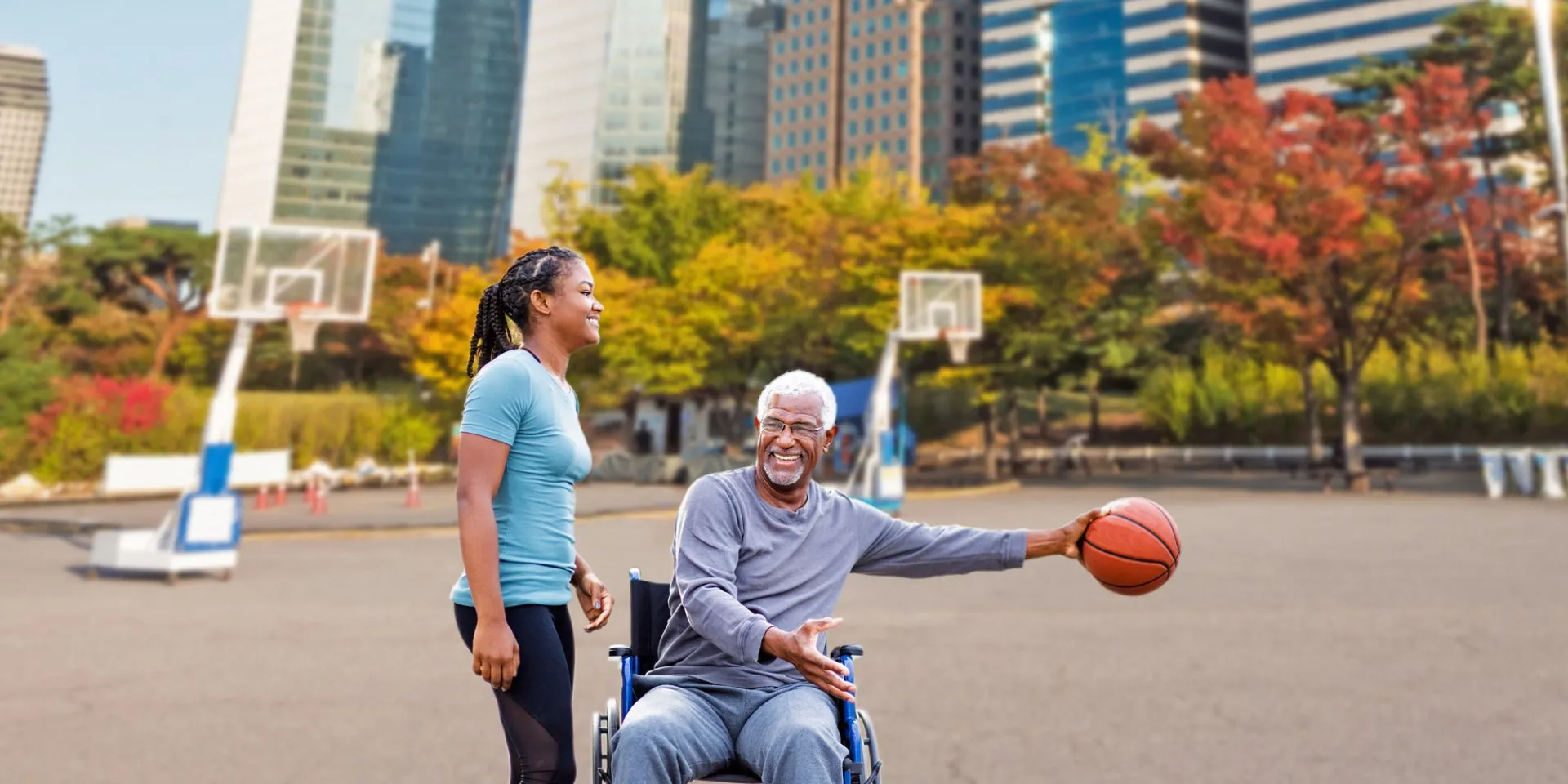 Older man in wheelchair holding basketball, smiling with standing woman on outdoor court.