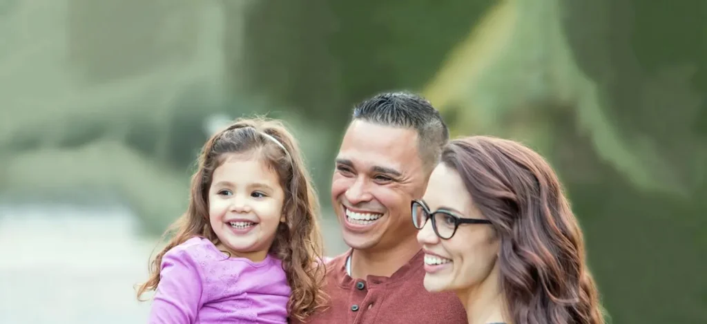 Smiling family of three outdoors, featuring a young girl in purple and adults with glasses and a red shirt.
